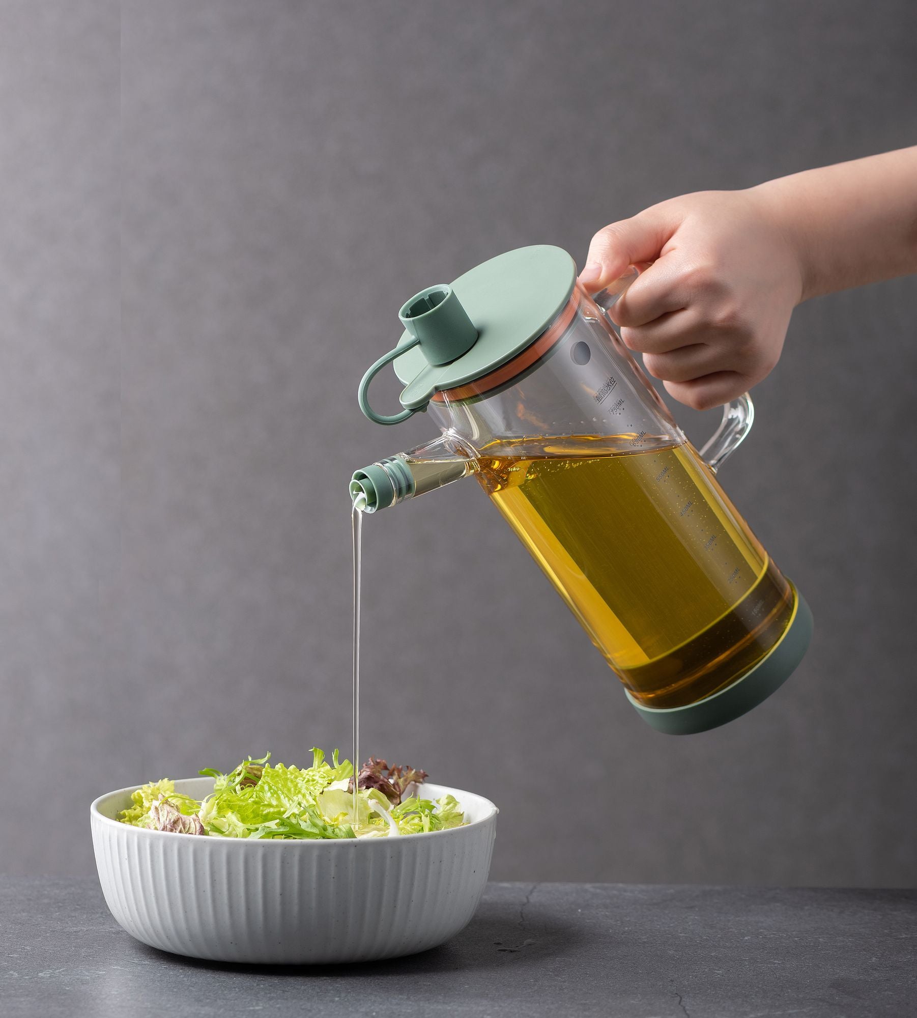 Huilier 75cl céladon versant de l'huile d'olive sur salade, main tenant le récipient en verre