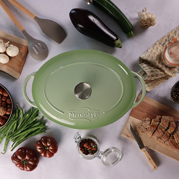 Cocotte ovale Colette 6,5L verte en fonte émaillée, vue du dessus entourée d'ingrédients frais et ustensiles de cuisine