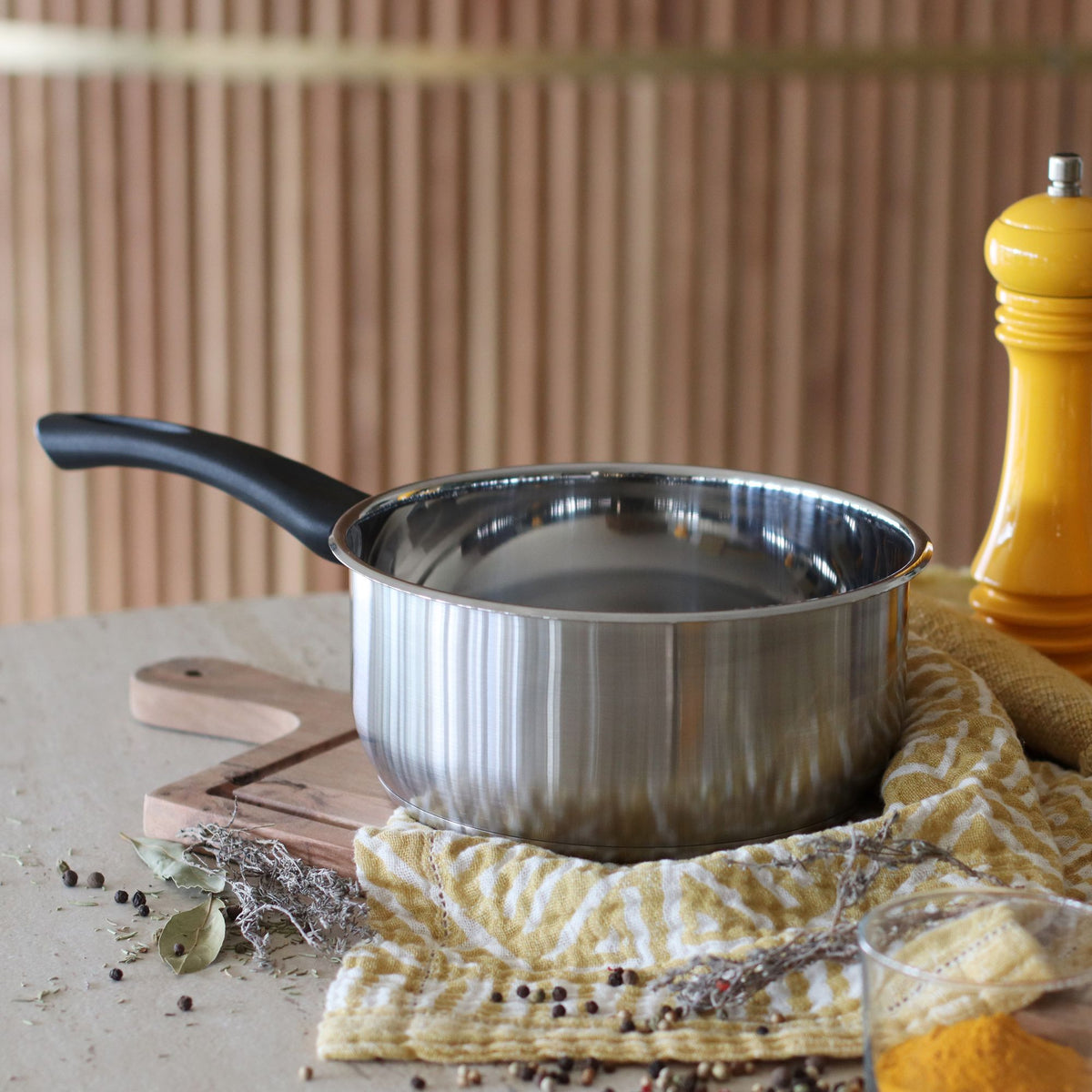 Casserole First 20 cm en acier inoxydable avec poignée noire, posée sur torchon jaune près d'un moulin à poivre