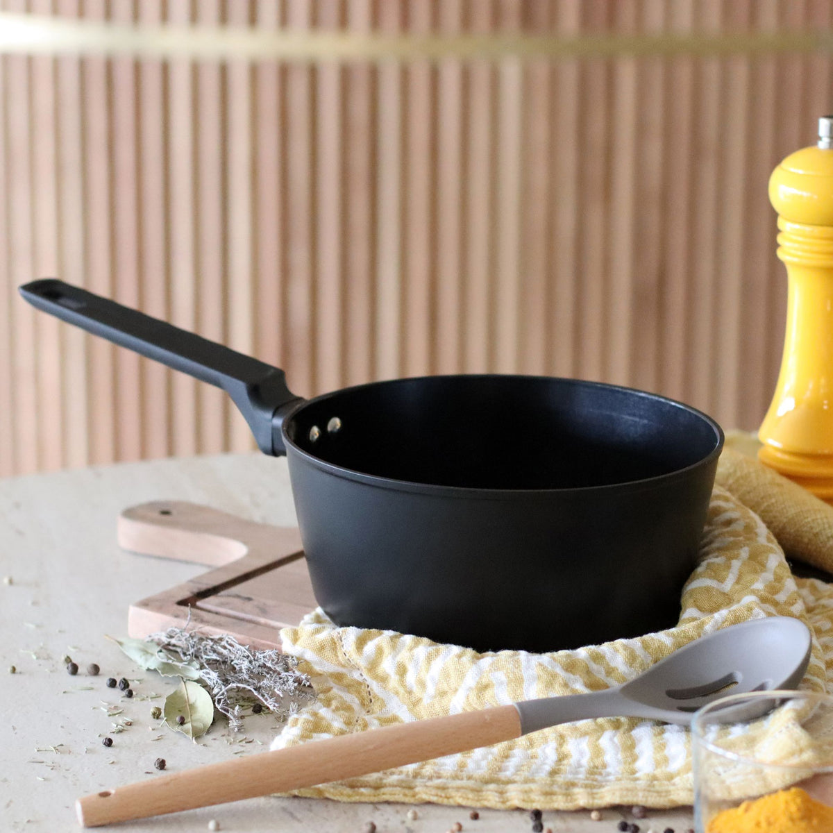 Casserole First 20 cm en aluminium noir sur torchon jaune avec ustensiles de cuisine et épices