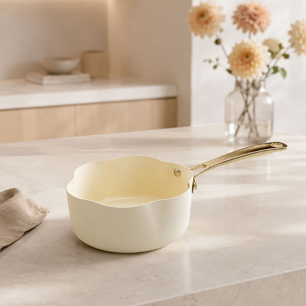 Casserole Dahlia 18 cm beige avec poignée dorée, posée sur plan de travail clair avec fleurs en arrière-plan