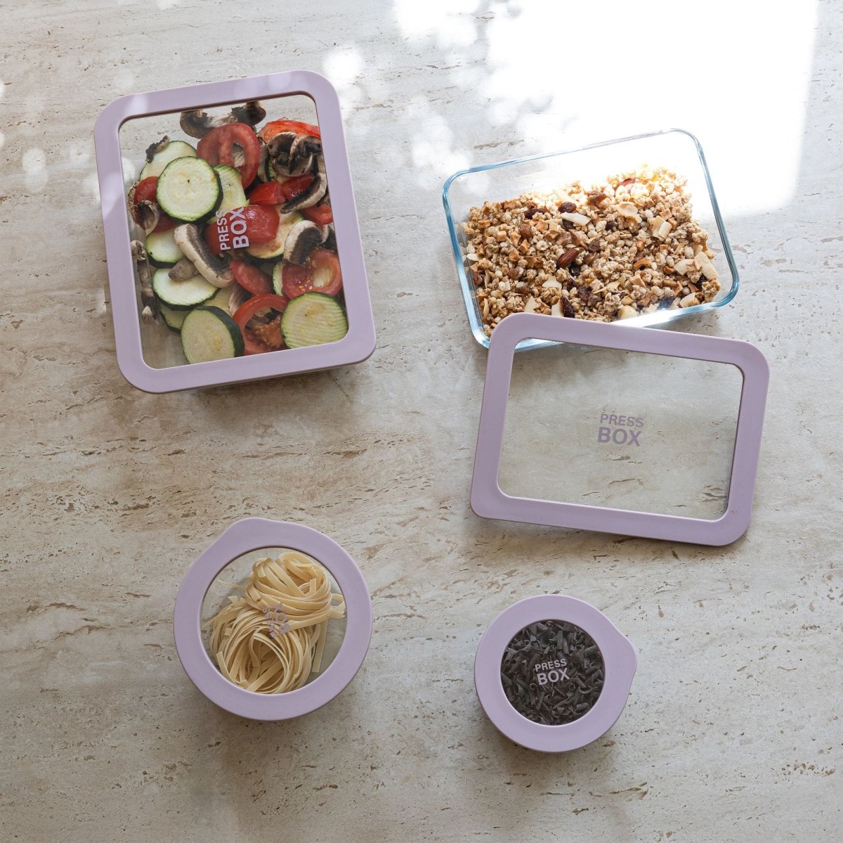 Vue d'ensemble de quatre boîtes hermétiques Pressbox en verre borosilicate avec cadres violets, contenant légumes, granola, pâtes et graines