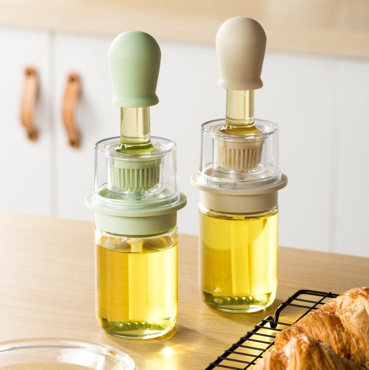 Deux bouteilles d'huile avec brosse intégrée en verre et silicone vert et beige, posées sur plan de travail bois