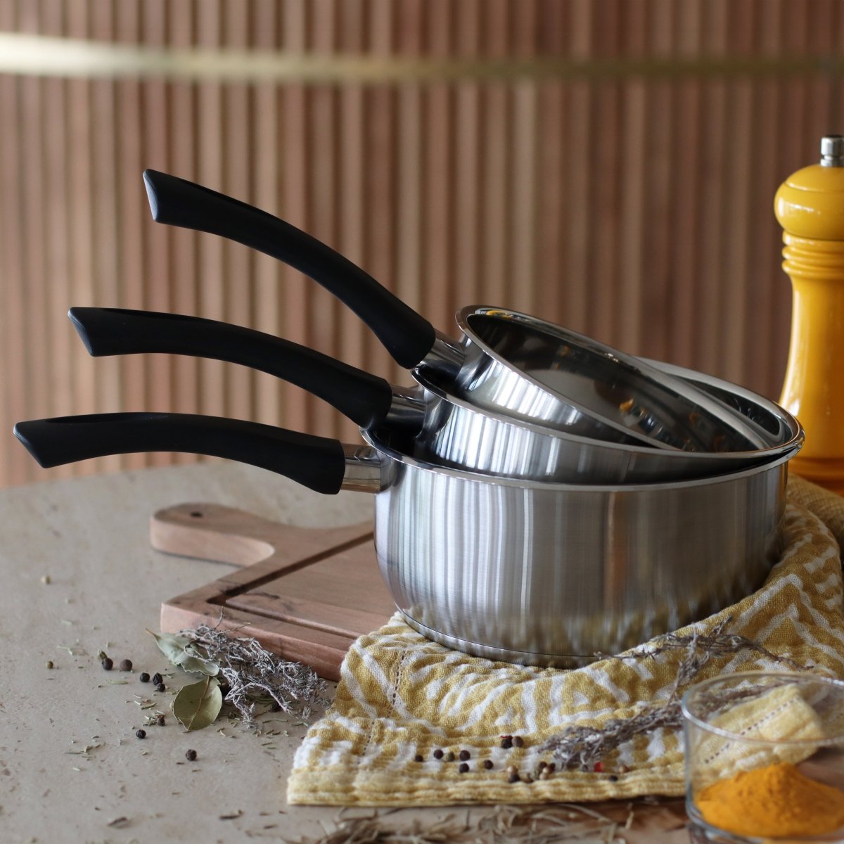 Set de 3 casseroles First en acier inoxydable avec poignées noires, posées sur plan de travail avec tissu jaune