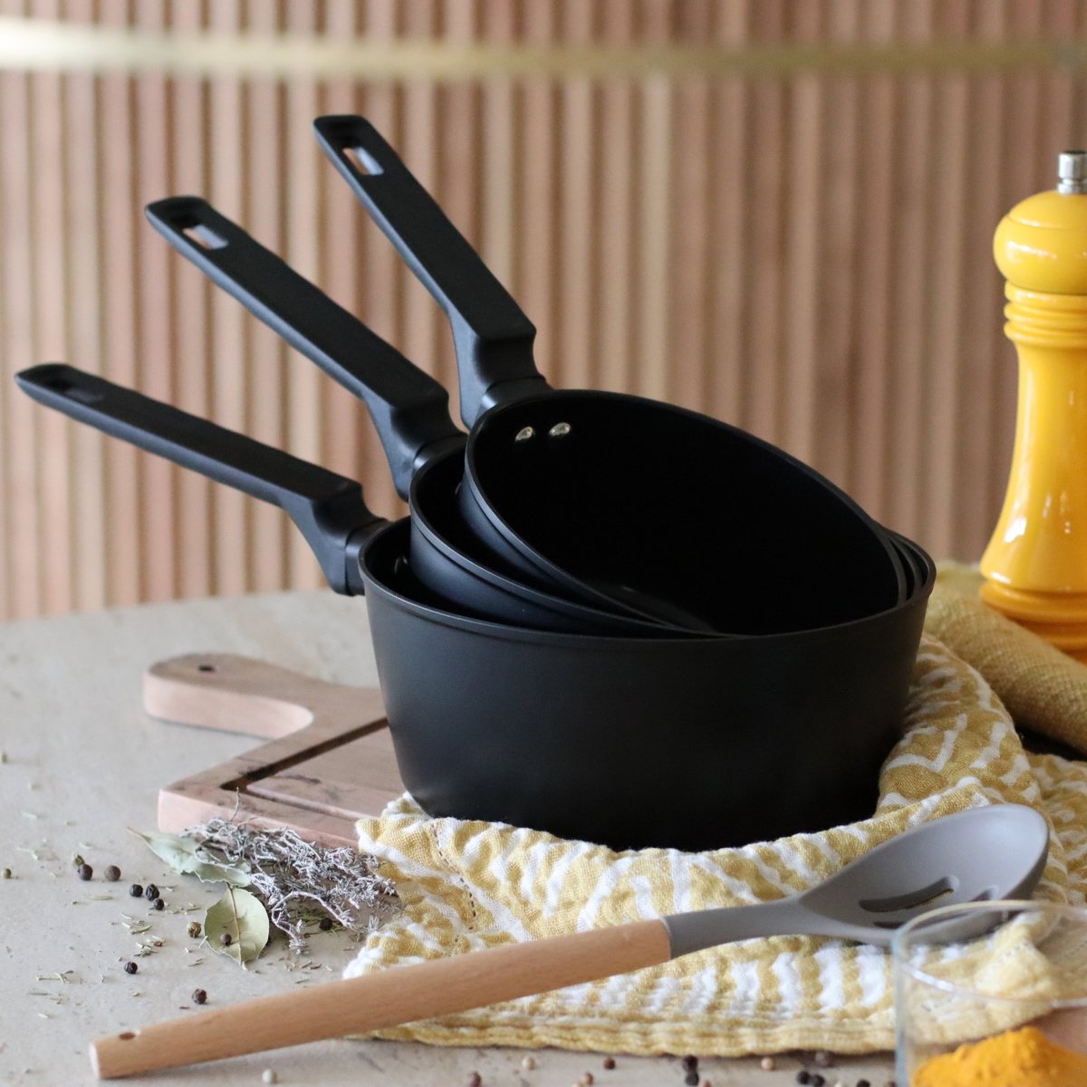Set de 3 casseroles First en aluminium noir empilées, décor culinaire avec torchon jaune et herbes aromatiques