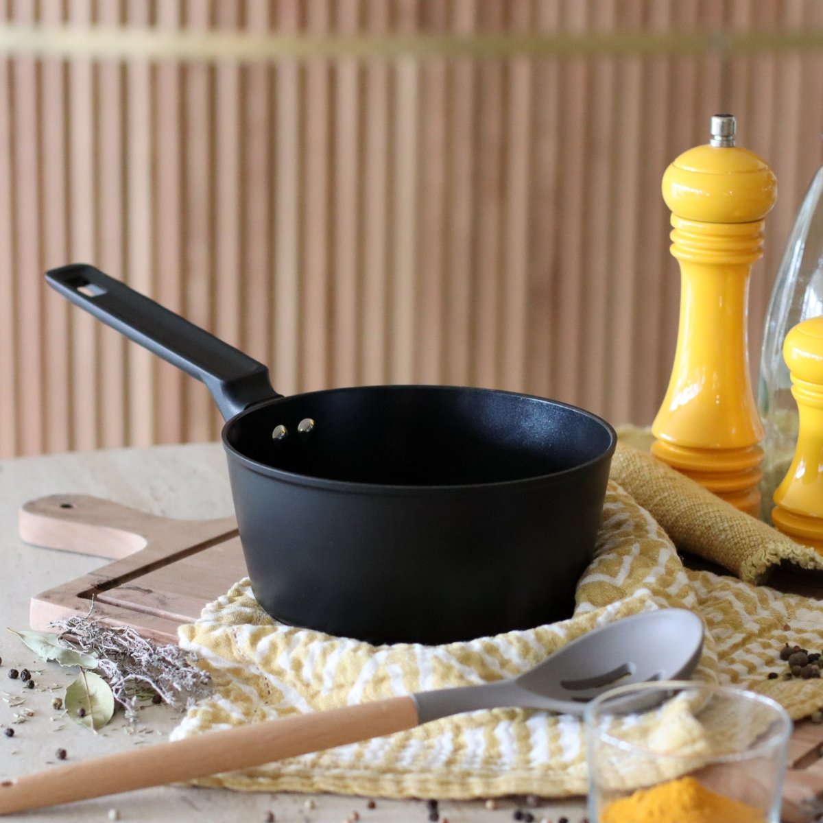 Casserole First 20 cm en aluminium noir posée sur torchon jaune avec ustensiles de cuisine en bois