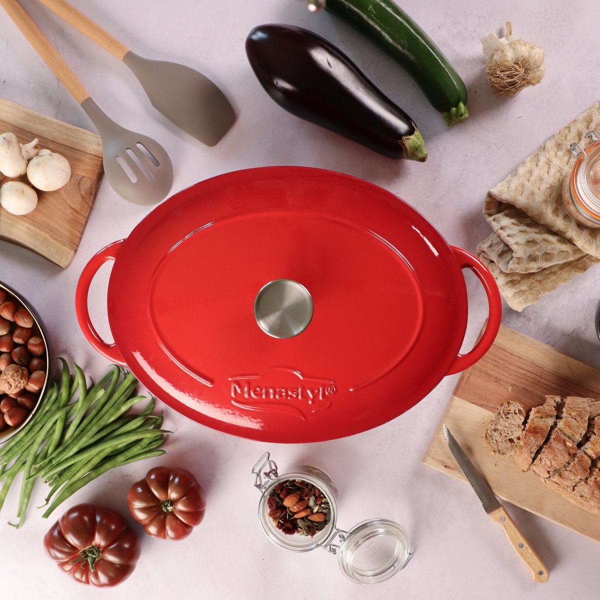 Cocotte ovale Colette 4L en fonte émaillée rouge, vue de dessus avec couvercle, entourée d'ingrédients frais