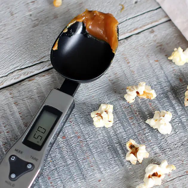 Cuillère à mesurer électronique affichant 50g avec caramel sur fond bois et pop-corn éparpillé