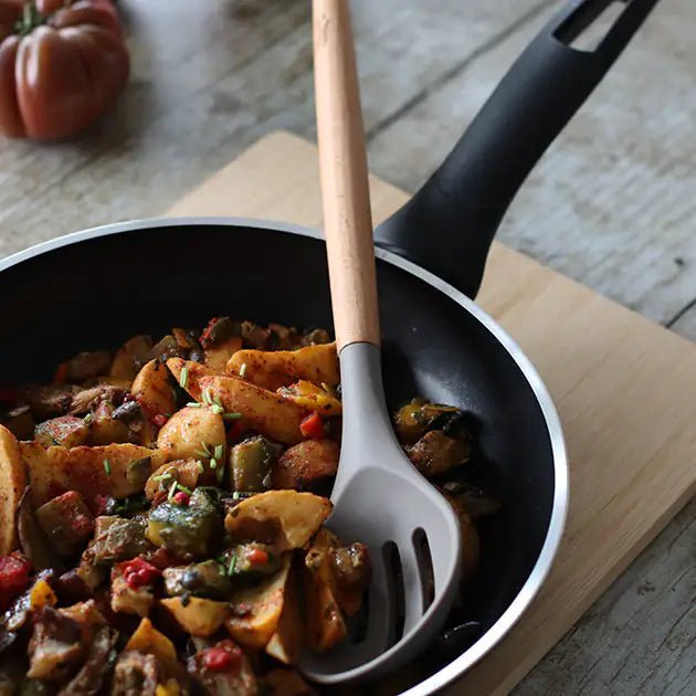 Cuillère ajourée Eliott en silicone gris et bois dans poêle avec légumes sautés, vue en action sur planche bois