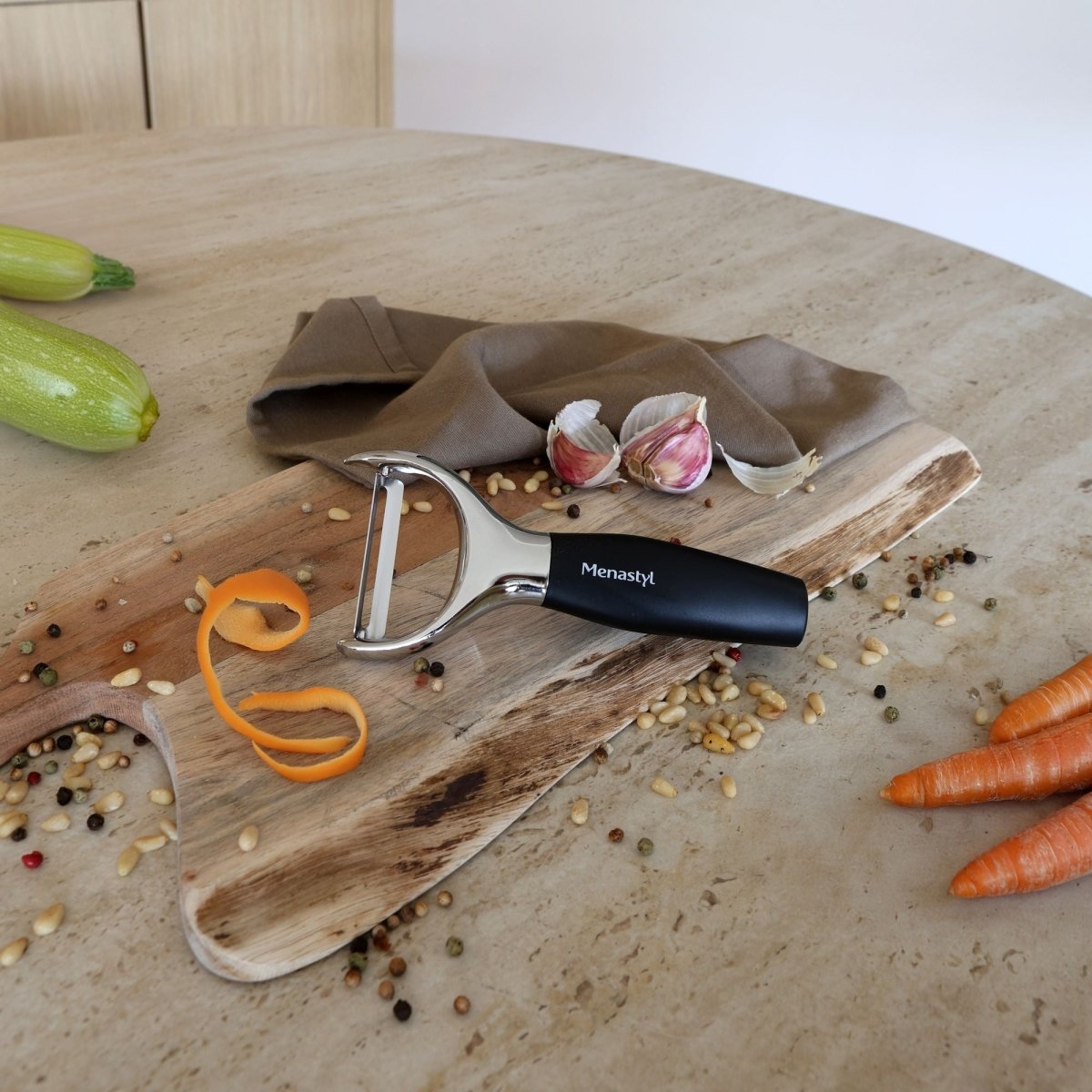 Épluche-légumes acier inoxydable à manche noir sur planche bois avec légumes, ail et épices