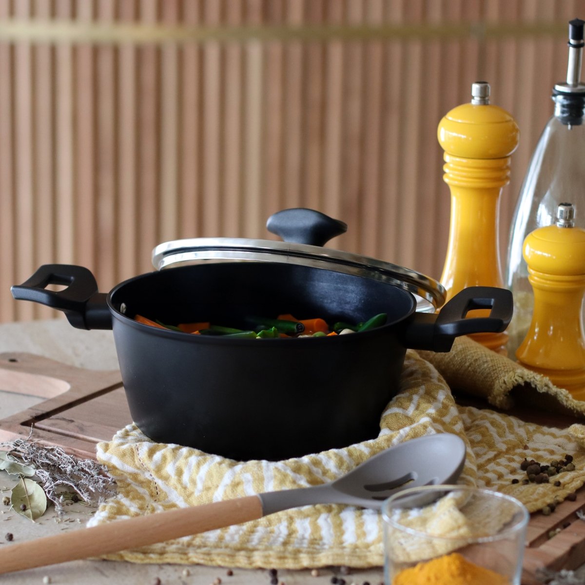 Faitout First 20 cm en aluminium noir sur table en bois avec ustensiles et condiments jaunes en arrière-plan