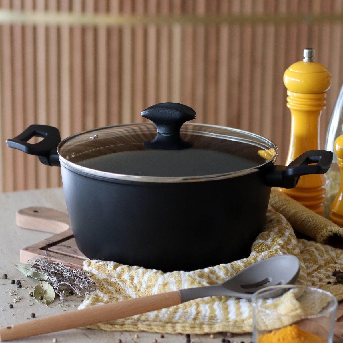 Faitout First 24 cm noir avec couvercle en verre, posé sur torchon rayé avec ustensiles de cuisine