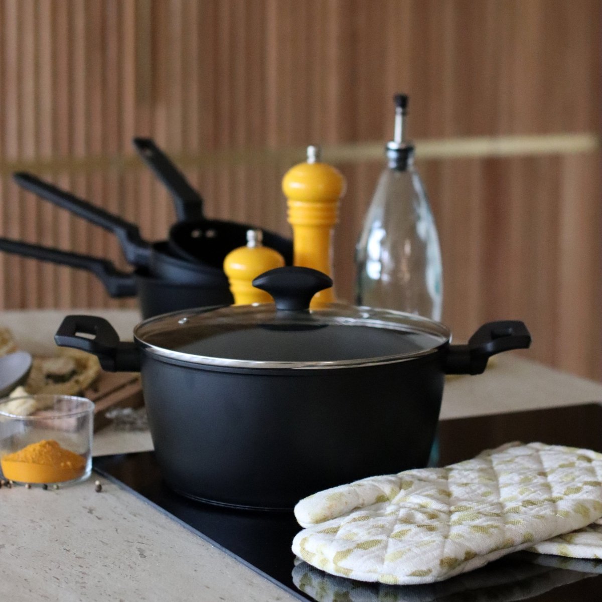 Faitout First 24cm noir avec couvercle verre, posé sur plan de travail avec ustensiles cuisine en arrière-plan