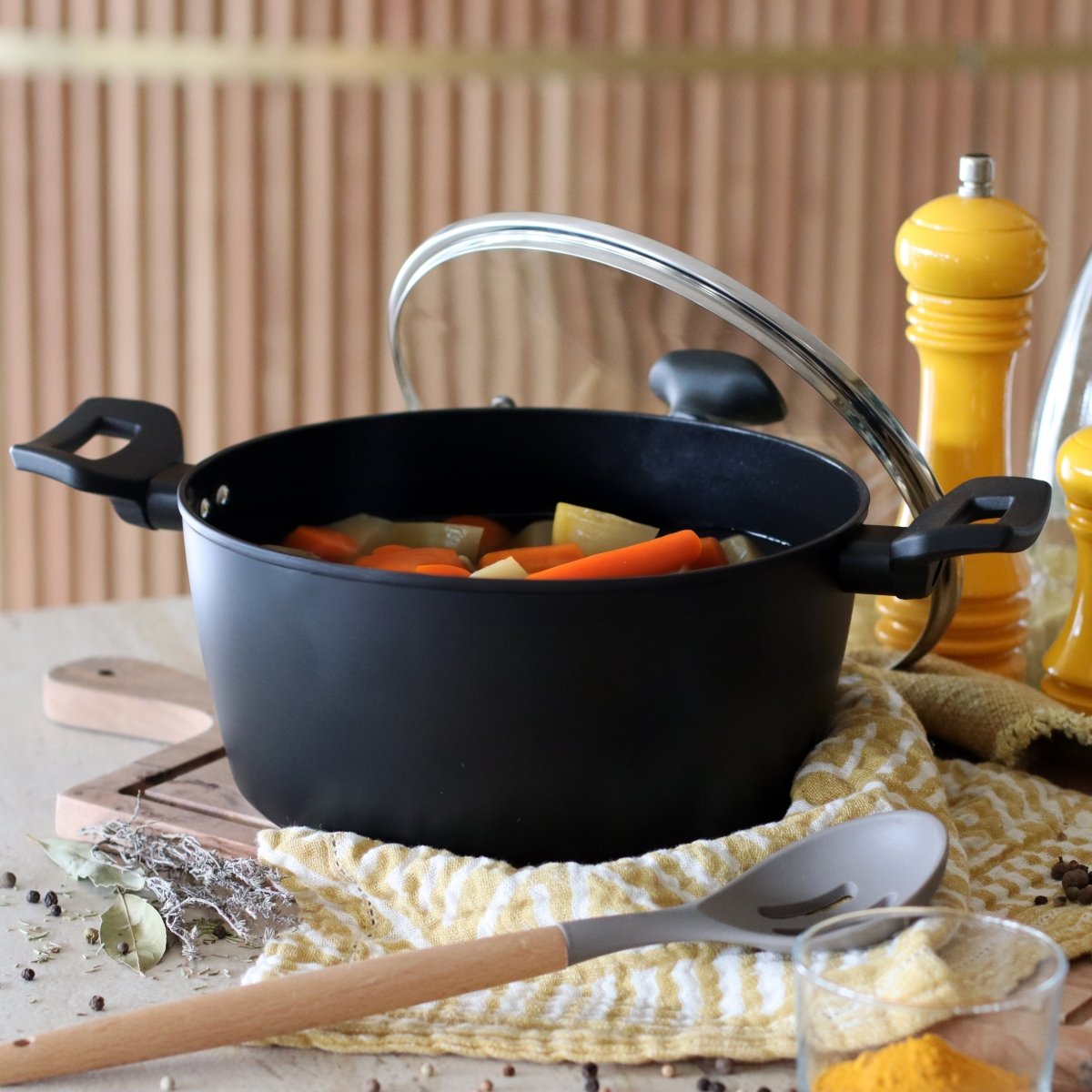 Faitout First 24 cm noir avec légumes, couvercle en verre et ustensiles de cuisine sur table