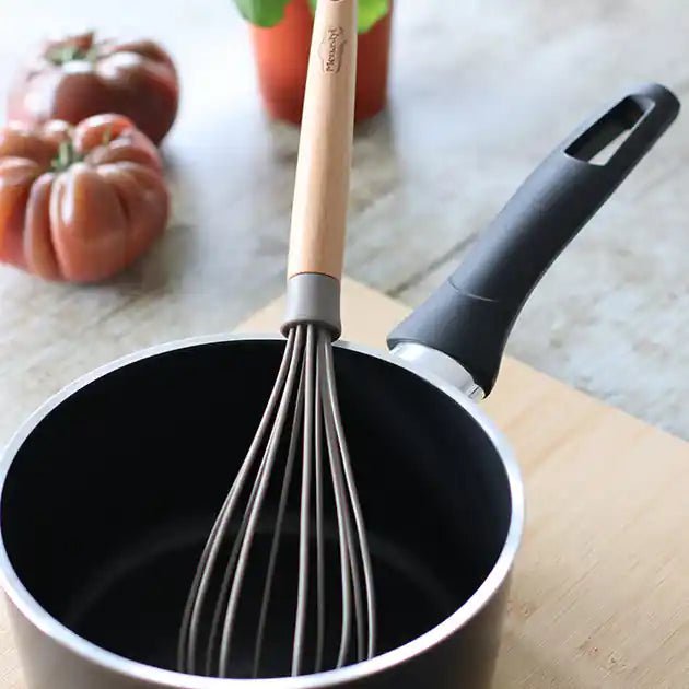 Fouet Eliott en silicone noir avec manche bois posé dans une casserole antiadhésive, sur plan de travail