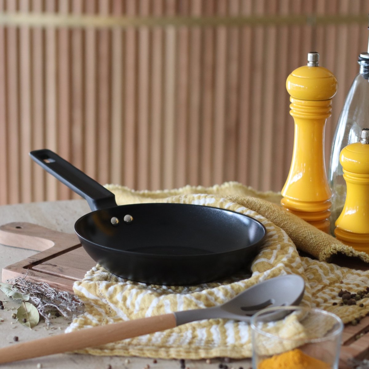 Poêle First 20 cm en aluminium noir posée sur torchon jaune avec ustensiles de cuisine en bois
