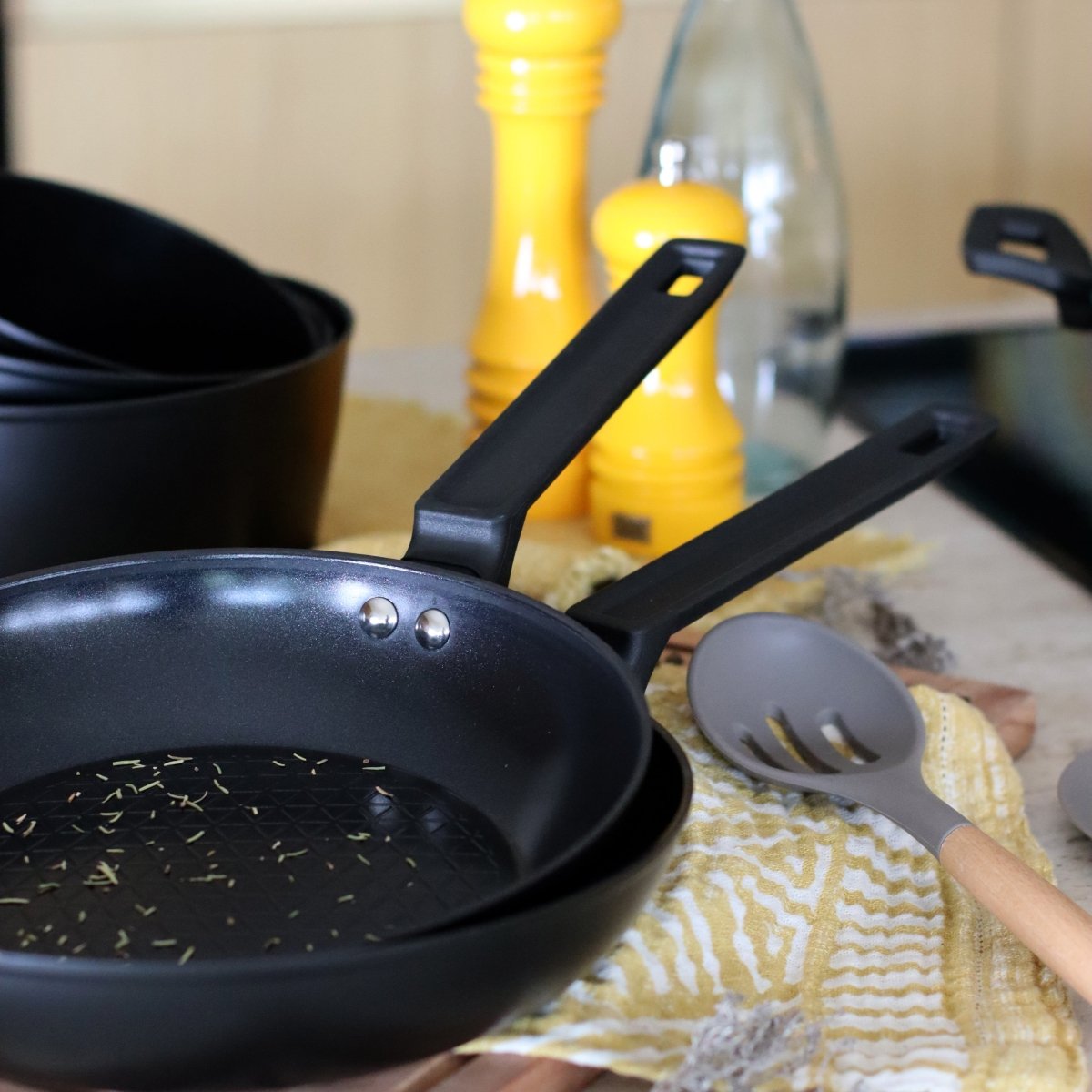 Poêle First 24cm en aluminium noir avec ustensiles de cuisine, setup complet sur torchon jaune