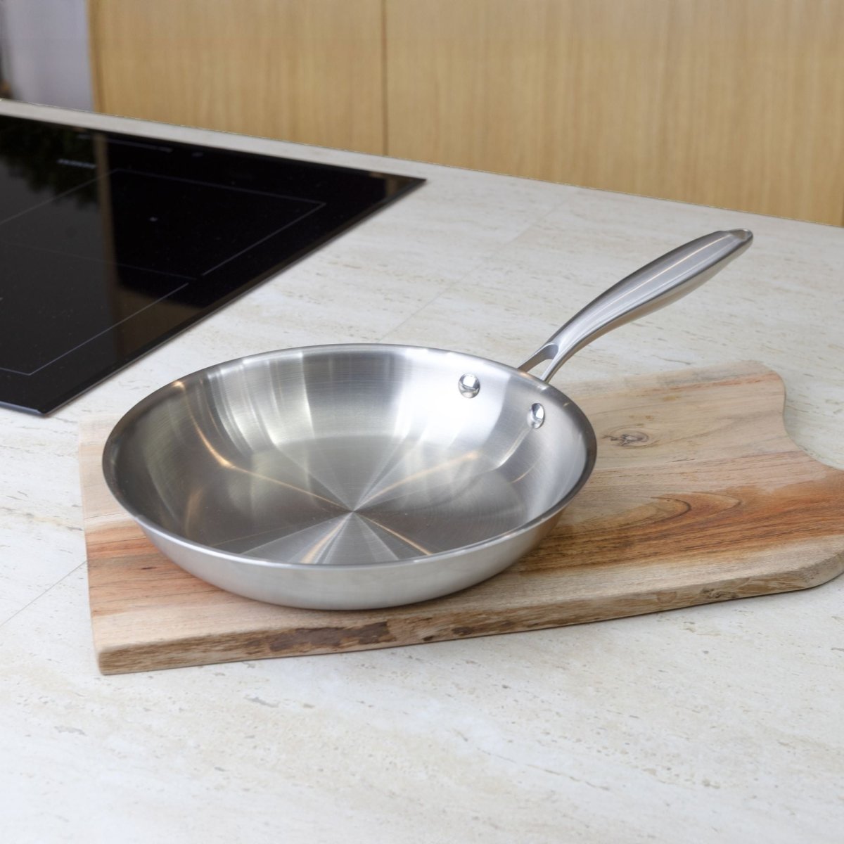 Poêle Trinox 24cm en acier inoxydable posée sur planche bois près d'une plaque de cuisson