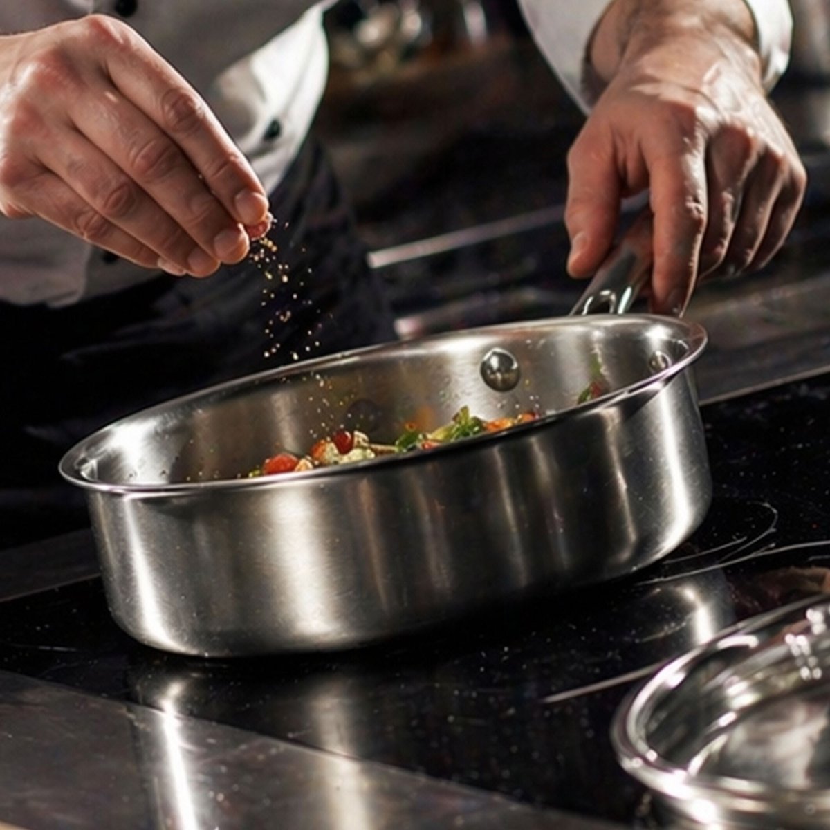 Chef cuisinant des légumes dans une sauteuse Triply en inox 24cm, assaisonnement en action sur plaque induction