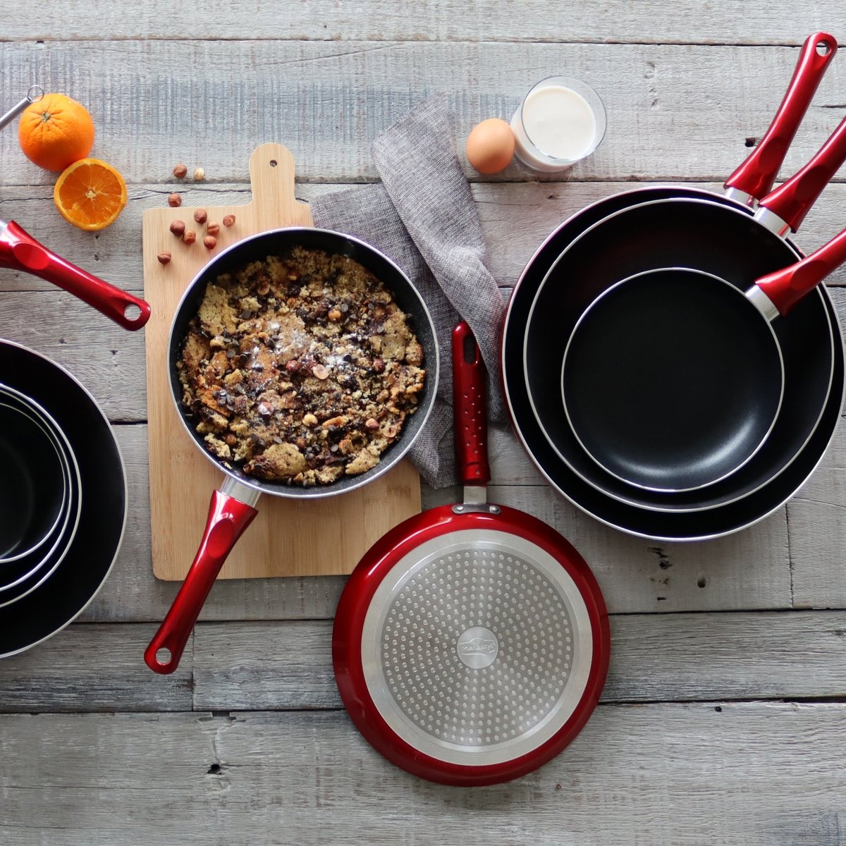 Set de 3 poêles aluminium rouge cerise 24/28/32 cm disposées sur table bois avec ingrédients cuisine