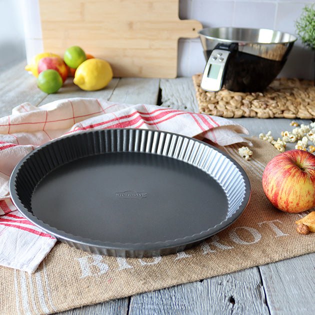 Moule à tarte Amandine 28 cm en acier carbone noir, cannelures antiadhésives, posé sur torchon avec pommes